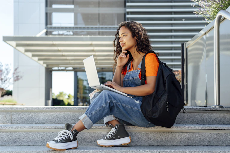 A woman sits on steps while holding a laptop and wearing a backpack.