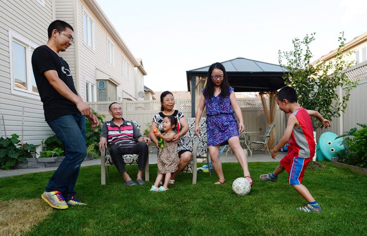 A family stands on the grass, two sit on lawn chairs, a soccer ball is on the ground. the woman is kicking it