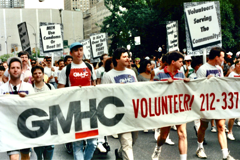 Group of people holding signs and marching in a parade behind a banner reading 'GMHC VOLUNTEER!'