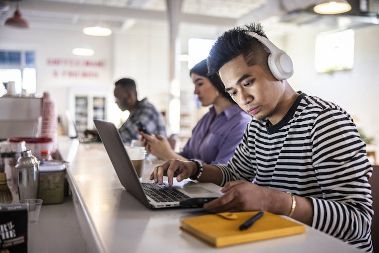 A man wearing headphones checks his phone while working on a laptop.