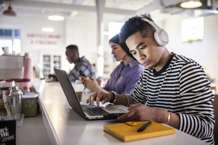 A man wearing headphones checks his phone while working on a laptop.