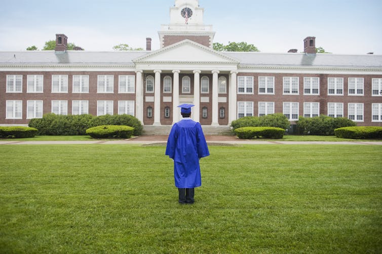 A student wearing a blue cap and gown stands on grass looking at a building.