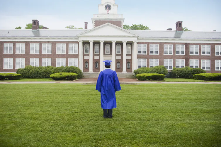 A student wearing a blue cap and gown stands on grass looking at a building.