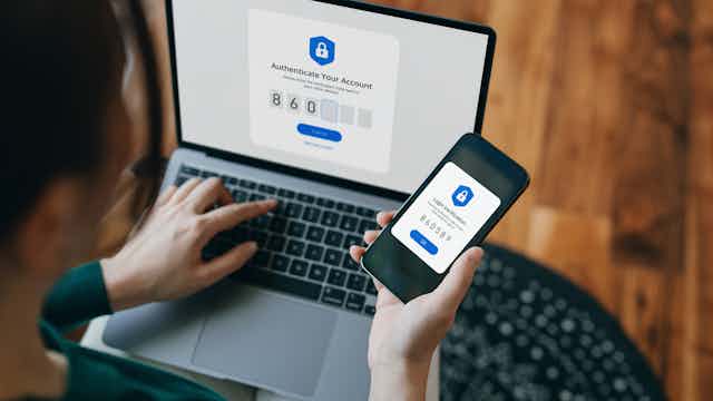 Right hand typing a number into a field on a laptop labelled authenticate your account, left hand holds the smartphone showing the authentication number