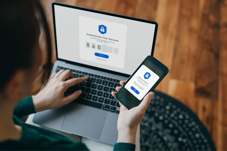 Right hand typing a number into a field on a laptop labelled authenticate your account, left hand holds the smartphone showing the authentication number