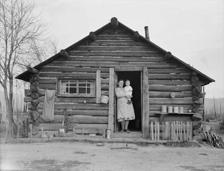 A mother holds her child in the front doorway of their log cabin home.