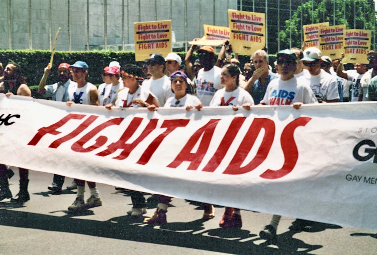 Group of people holding signs reading 'Fight to Live, Fight to Love, Fight AIDS' and marching in a parade behind a banner reading 'FIGHT AIDS'