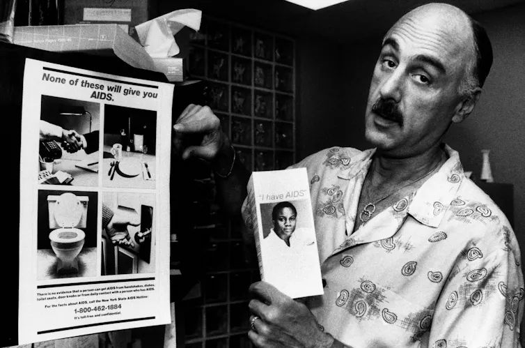 Person holding a pamphlet reading 'I have AIDS,' beside a poster of a handshake, a toilet, a doorknob and an empty plate, reading 'None of these will give you AIDS.'