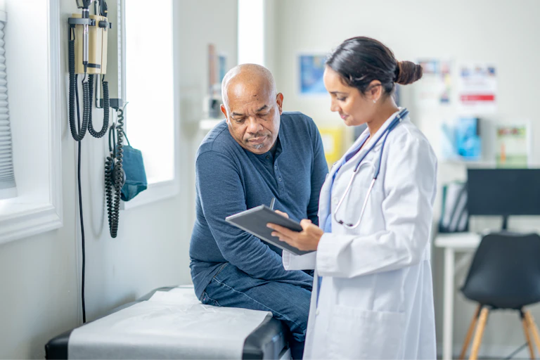 A older man sits up on an exam table during a routine check-up. His female doctor is holding out a tablet as they review some recent test results together.