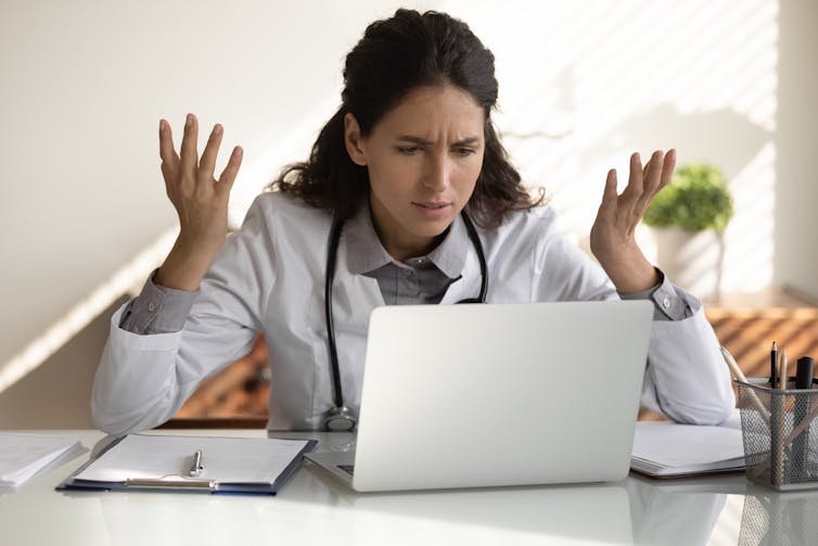 A female doctor looking at a computer screen throws up her hands in frustration.