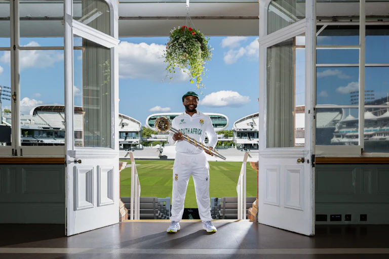 An African man in white cricket outfit stands between open white wooden doors holding a large silver and gold sceptre and smiling, behind him a cricket field.