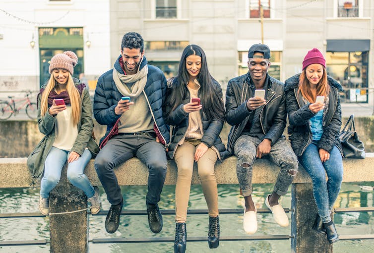 group of five young people sat by river, all looking at phones