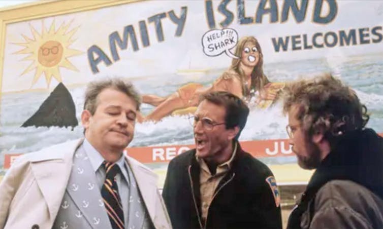 Three men arguing in front of a billboard for a seaside holiday town called Amity Island, with a shark fin drawn on it.