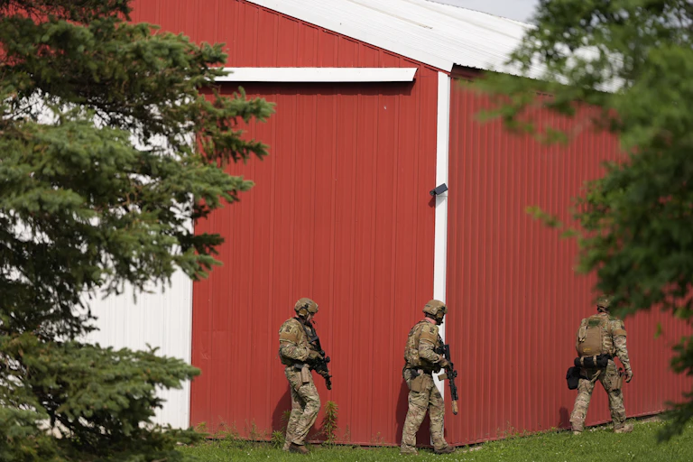 Three police officers, wearing helmets and carrying guns, walk near a home with a red outer wall.