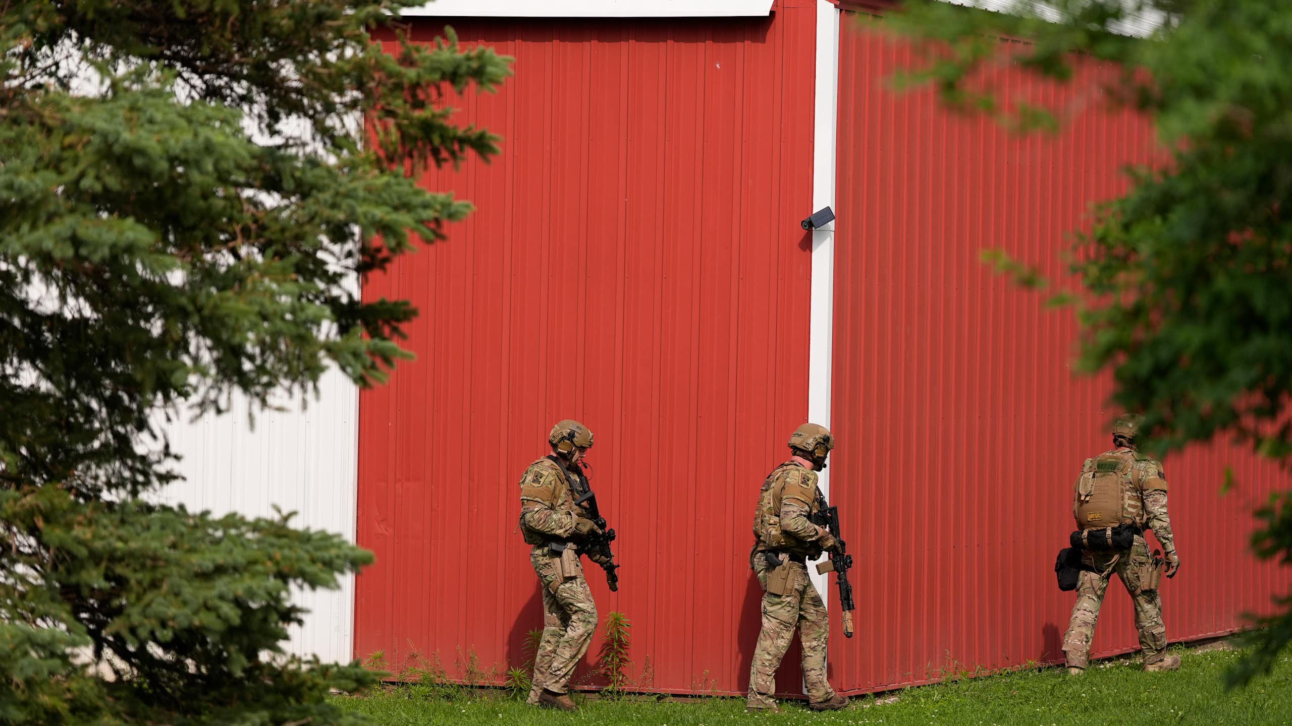 Three police officers, wearing helmets and carrying guns, walk near a home with a red outer wall.