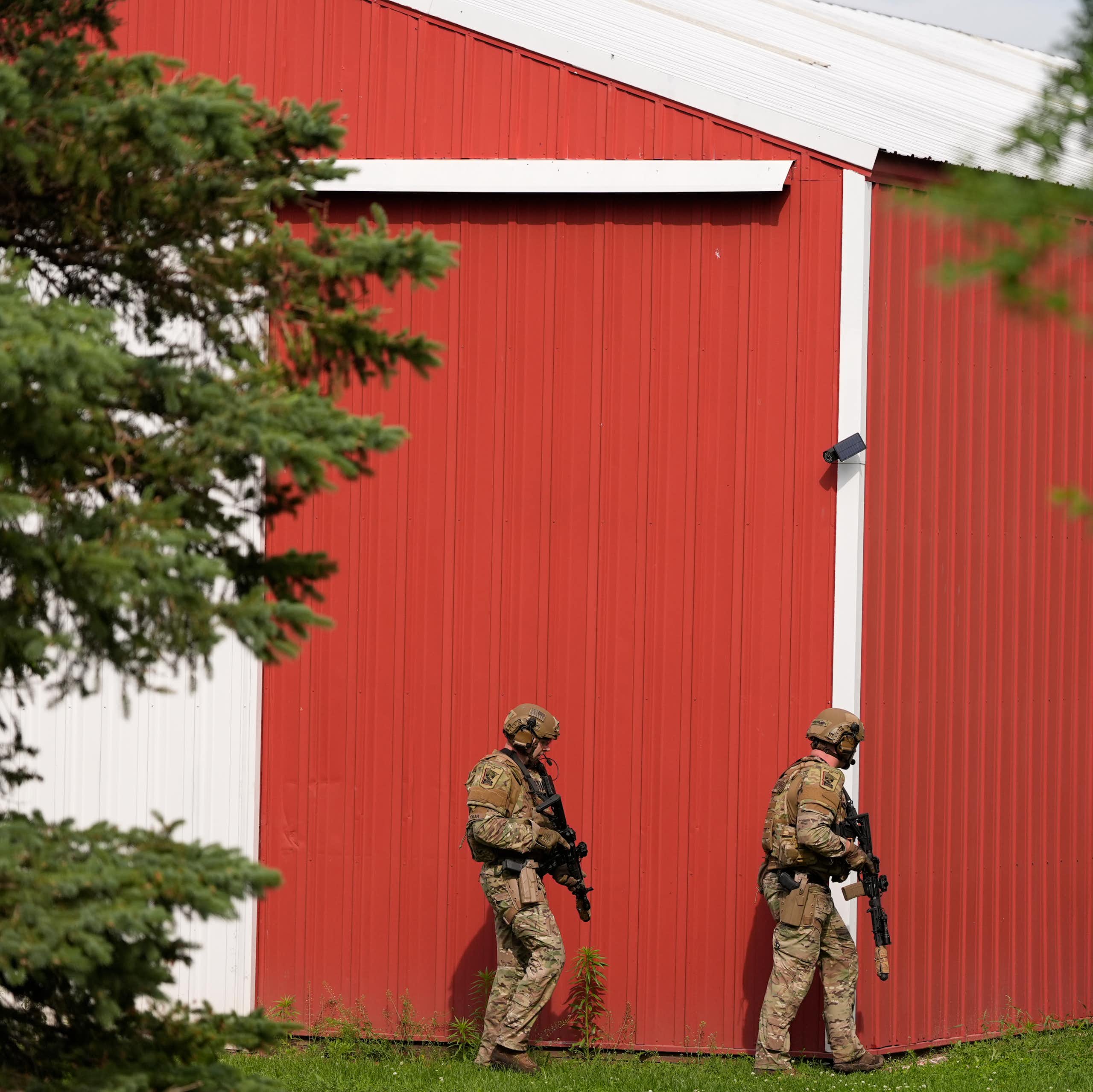 Three police officers, wearing helmets and carrying guns, walk near a home with a red outer wall.