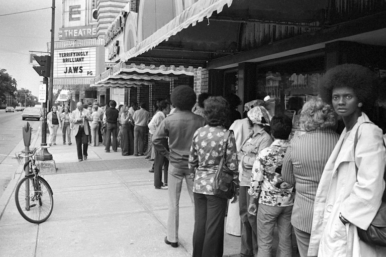 A long line of people stand outside of a movie theater.