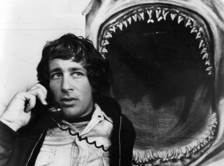 Young man with shoulder-length hair speaks on the phone in front of an image of a shark with its mouth open.