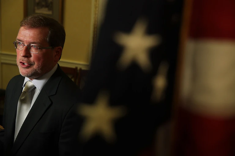 A man dressed in a suit speaks behind a U.S. flag.