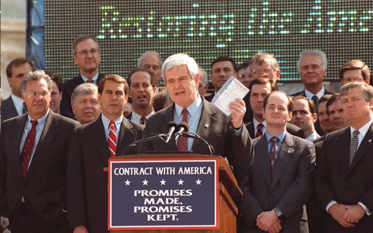 A man dressed in a suit and holding a book speaks in front on an outdoor lectern as other men stand behind him.