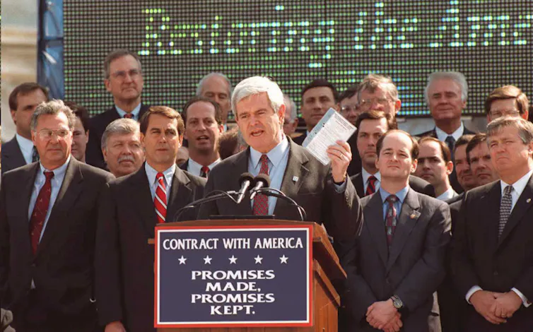 A man dressed in a suit and holding a book speaks in front on an outdoor lectern as other men stand behind him.
