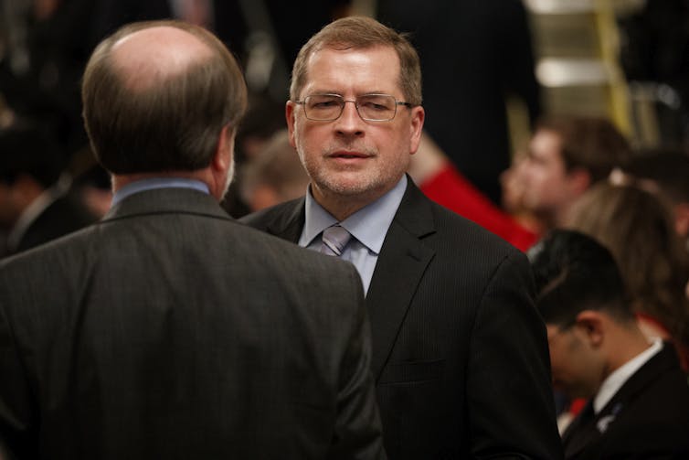 A man in a suit and wearing glasses looks down, as another man, his back to the camera, stands in front of him.