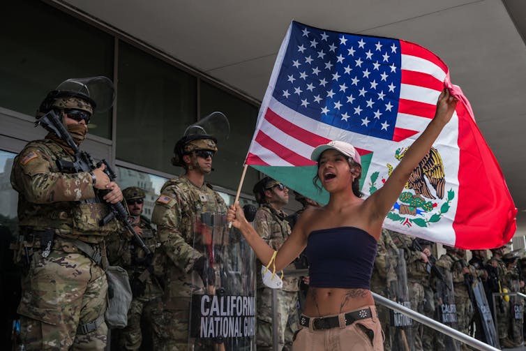 A woman carrying a Mexico-U.S. flag walks in front of soldiers.