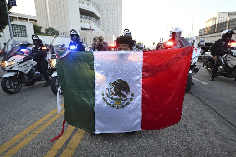 A man holds a Mexican flag in front of several police officers on motorcycles.