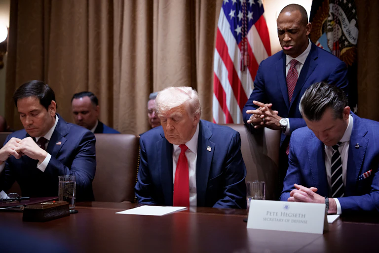 Three men in navy blue suits sit with their heads bowed in prayer, while one man stands behind.