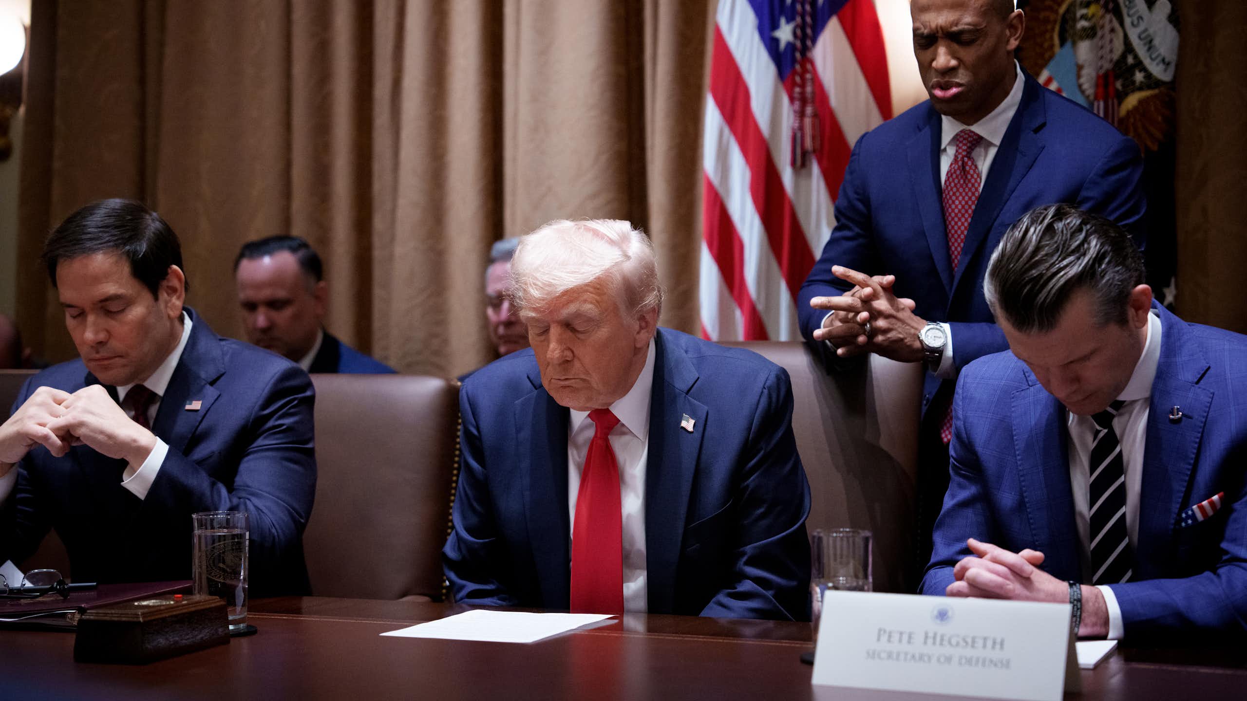 Three men in navy blue suits sit with their heads bowed in prayer, while one man stands behind.