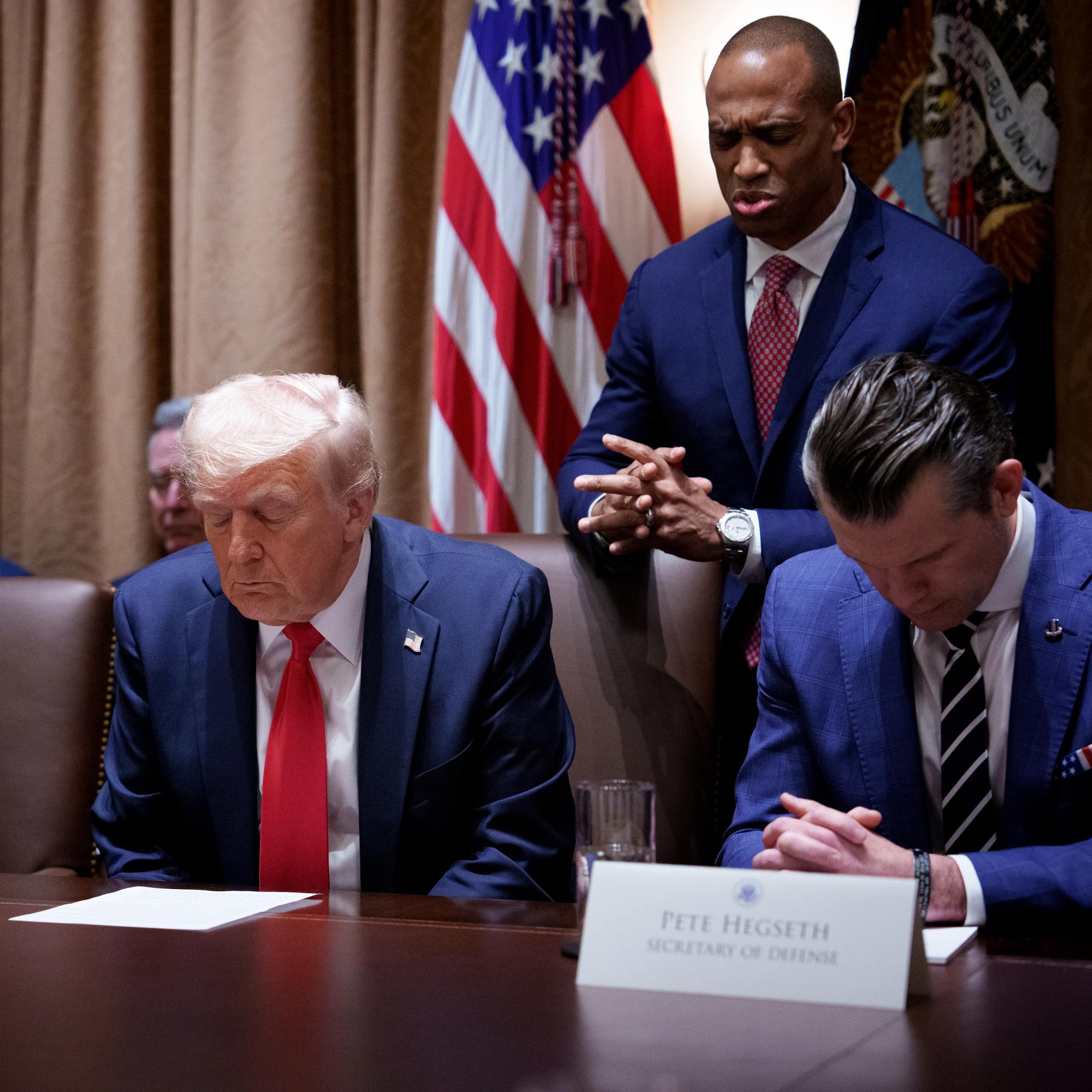 Three men in navy blue suits sit with their heads bowed in prayer, while one man stands behind.