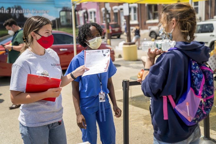 Three women wearing face masks, with a dressed in blue medical scrubs, speak together on a sidewalk lined with row houses
