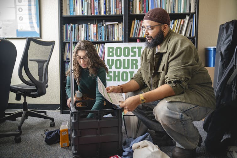 A man and a woman draw papers from a foldable cart while a poster behind them reads