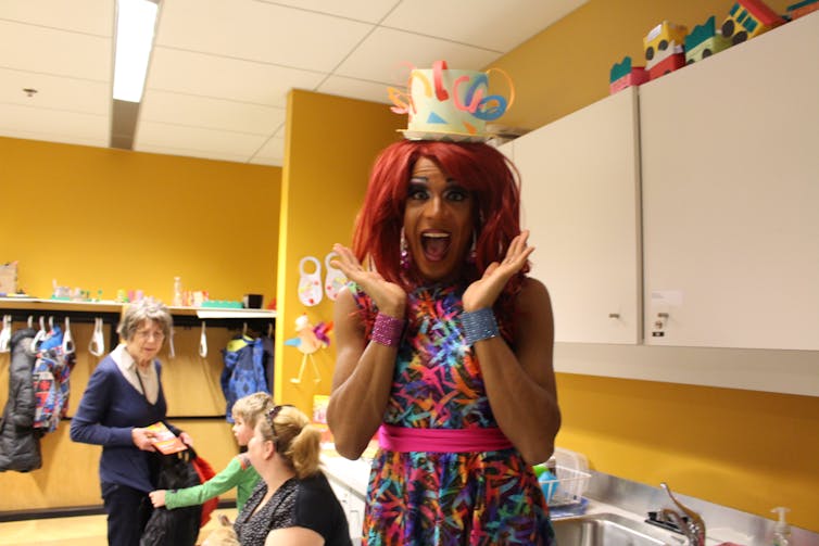 A smiling person with outstretched and raised hands in a multicoloured dress with what looks like a paper teacup on their head.