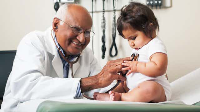 An elderly doctor with glasses places a stethoscope to a baby in a doctor's office.