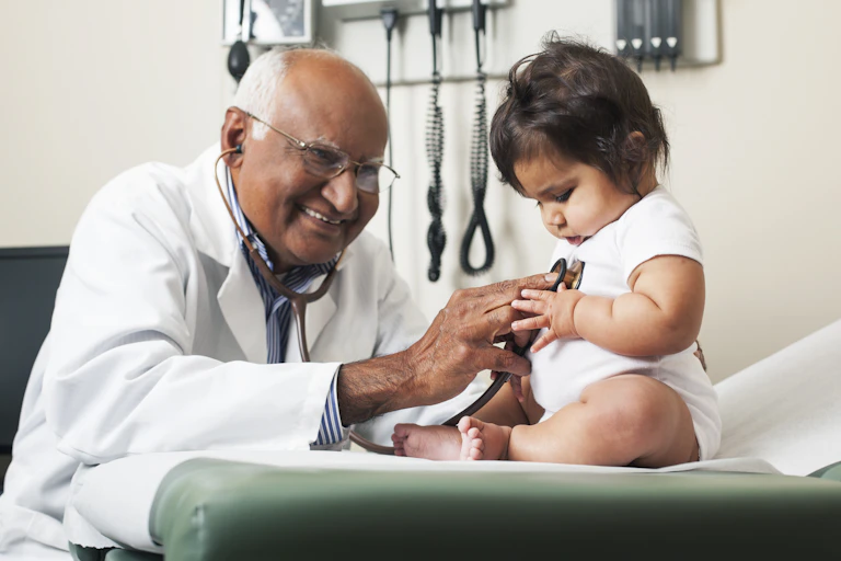 An elderly doctor with glasses places a stethoscope to a baby in a doctor's office.