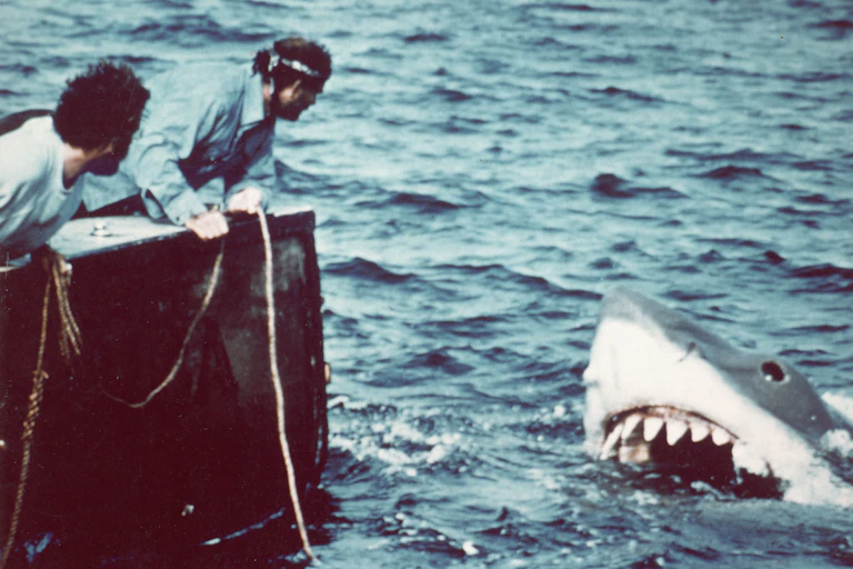 two men lean over side of a boat toward what is the snout of a mechanical shark in the water