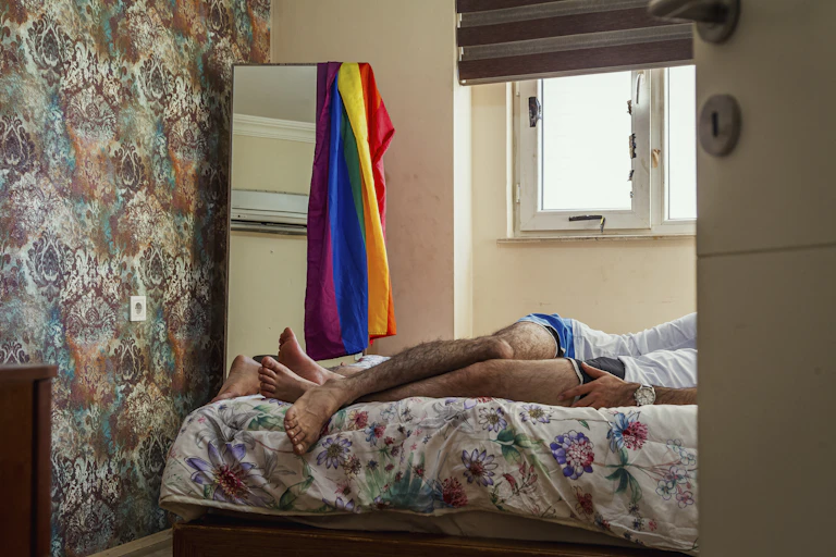 cropped image of a bedroom with the legs of two men visible and pride flag in the background.