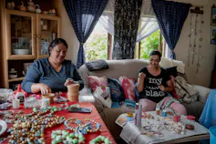 Two women sit inside working at tables covered with jewellery made from shells