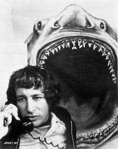 A young nerdy looking guy on a landline phone in front of a poster of a great white shark, its jaws wide open above his head.