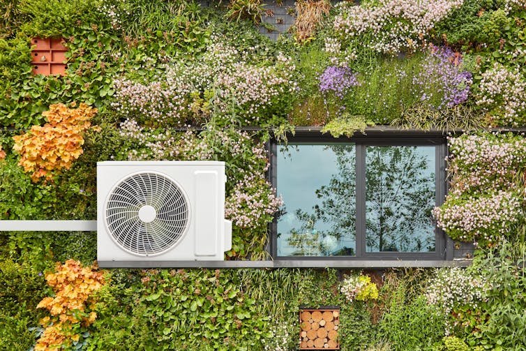 An air-source heat pump on a house covered in plants.