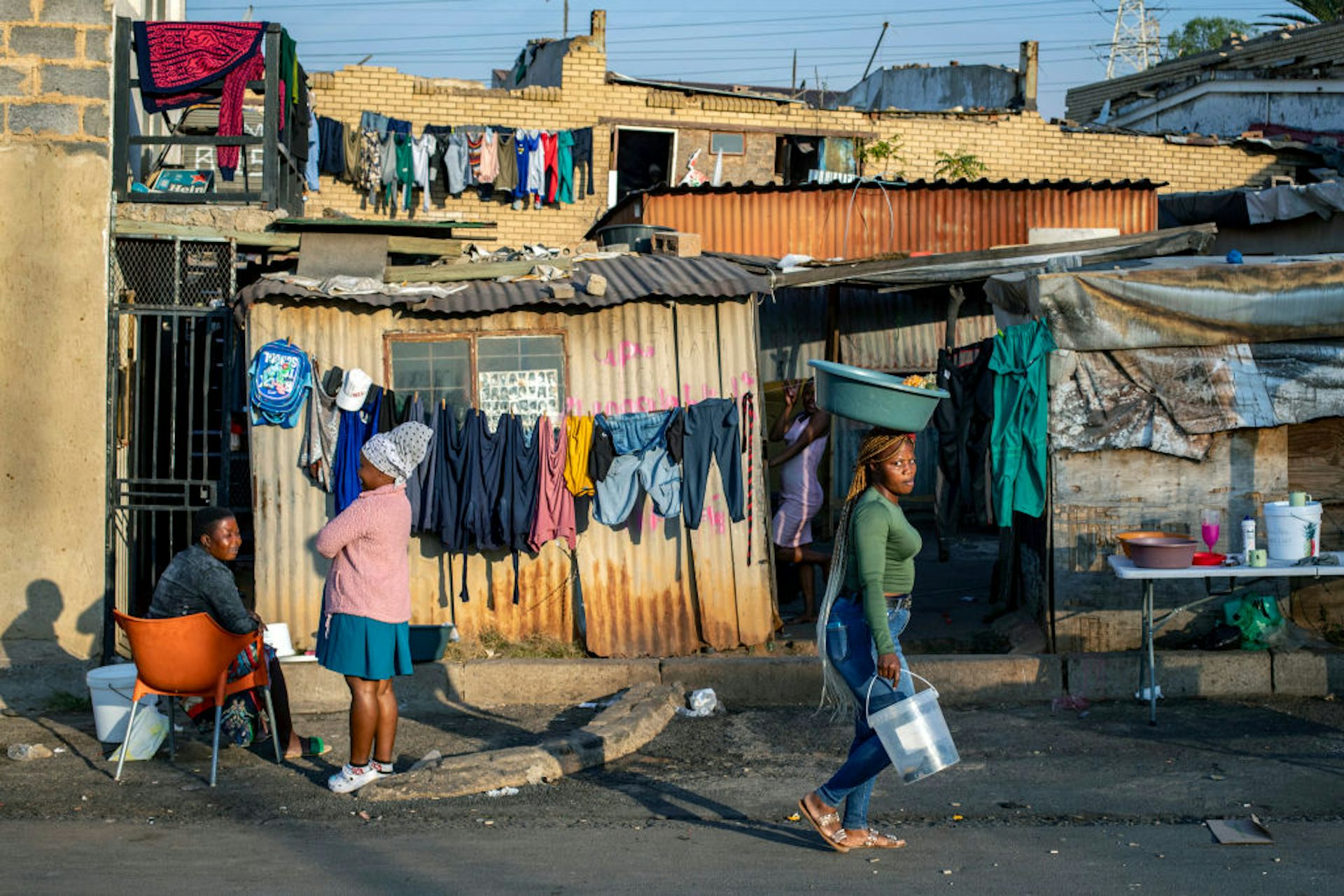 Shanty scene with washing lines, woman walking carrying a bucket and a basin, other women sitting and standing outside a corrugated iron shack