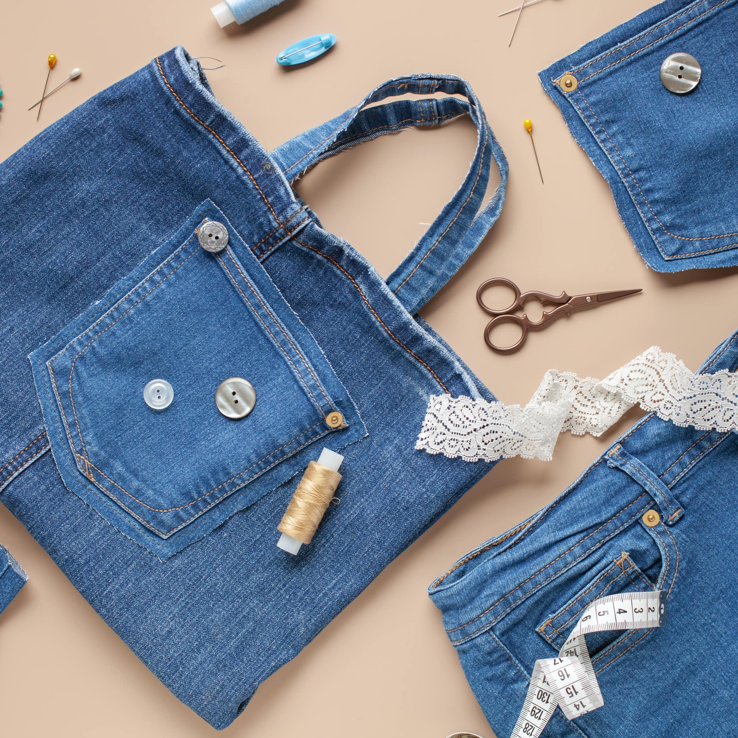 A handbag made of blue denim, with what appears to be the pocket from a pair of jeans sewn onto it, is on a surface with other pieces of denim, a lace-like material, and scissors and thread.