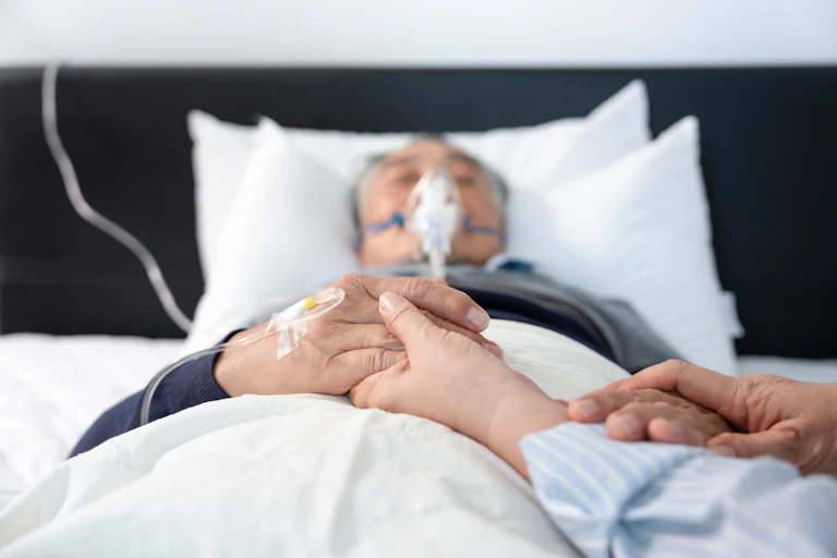 Un homme portant un masque est alité. Une personne lui tient les mains.