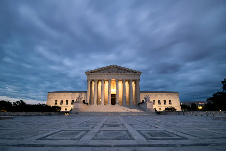 A very large white, columned building with a plaza in front and ominous clouds behind it.