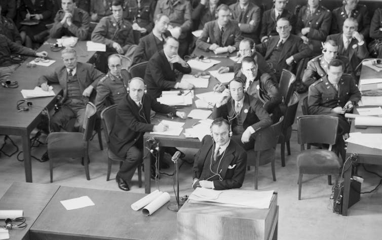 A meeting with many people seated at long tables that are covered with papers.