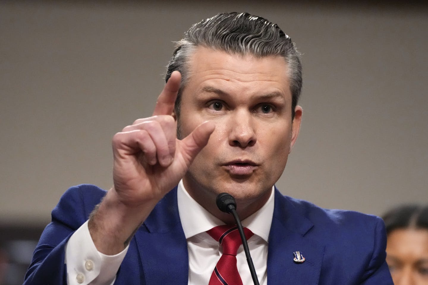 A man in a navy blue suit and red tie looks ahead while gesturing with his finger.