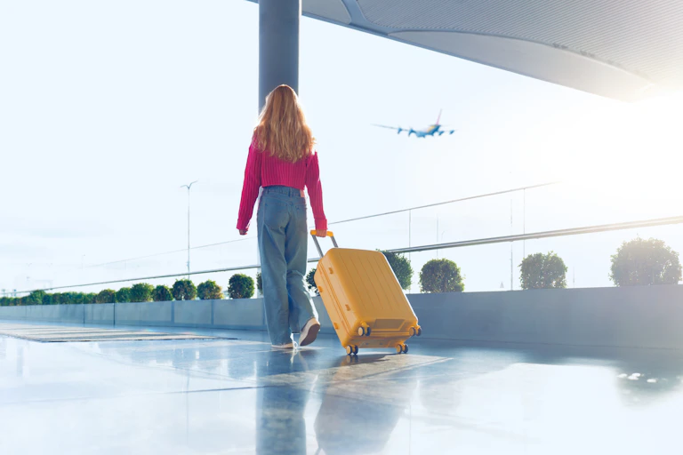 Una chica pelirroja camina por el exterior de un aeropuerto con una maleta en la mano mientras un avión vuela por el cielo de fondo