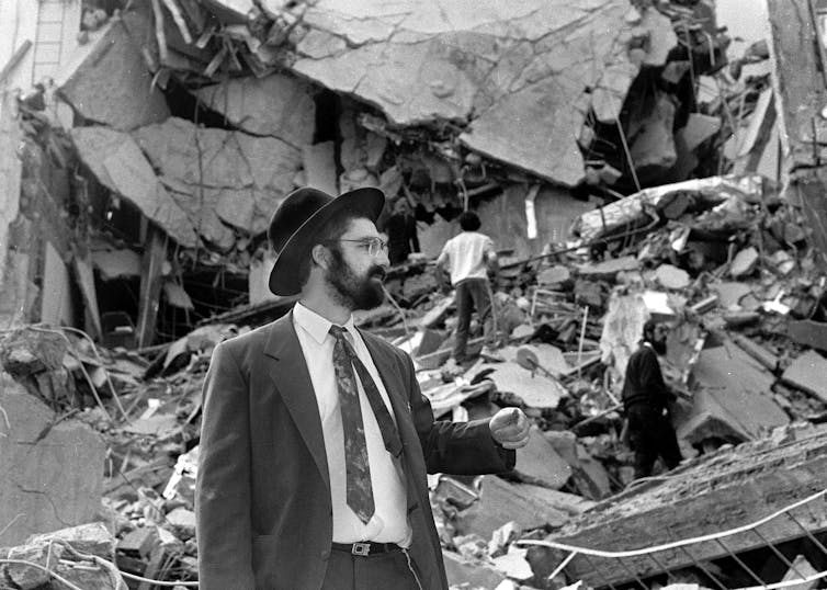 A man in a suit and hat walks through rubble.