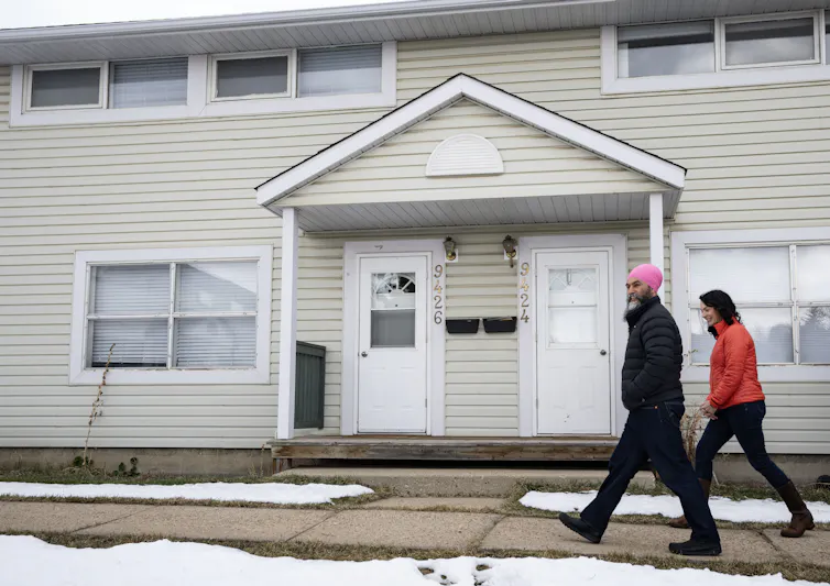 Two people, one a man wearing a bright pink turban, walk along a snowy residential street.